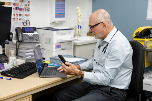 Doctor in office looking at phone and laptop - Australian Stock Image
