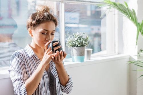 Early twenties woman enjoying a mug of coffee by a warehouse apartment window - Australian Stock Image