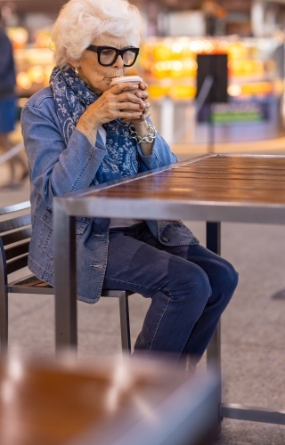 elderly lady wearing denim sitting at cafe table alone and sipping coffee in a disposable cup - Australian Stock Image