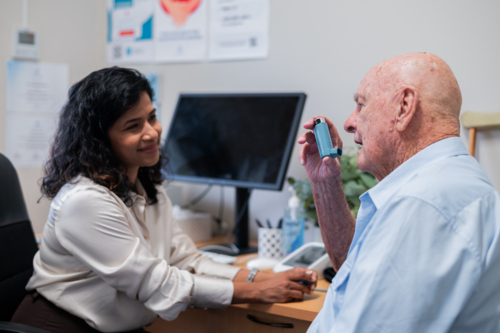 Elderly man holding an inhaler while seated facing healthcare practitioner - Australian Stock Image