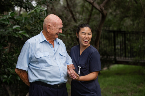Elderly man in light blue short-sleeved shirt holding hands with female caregiver in navy scrubs - Australian Stock Image