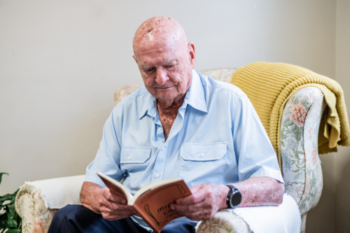 Elderly man sitting comfortably on a patterned armchair reading a book indoors - Australian Stock Image
