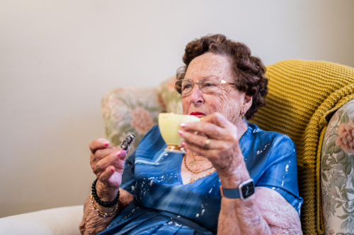 Elderly woman enjoying a cup of tea and a piece of lamington cake while seated on patterned armchair - Australian Stock Image