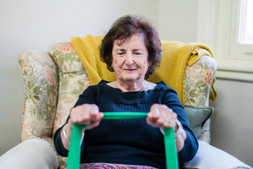 Elderly woman holding a green resistant band while seated on an armchair for strength training - Australian Stock Image