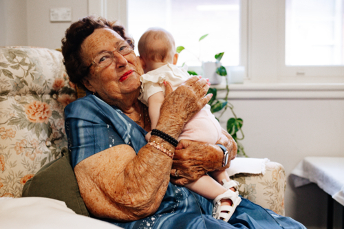Elderly woman holding baby girl while seated on floral patterned armchair - Australian Stock Image