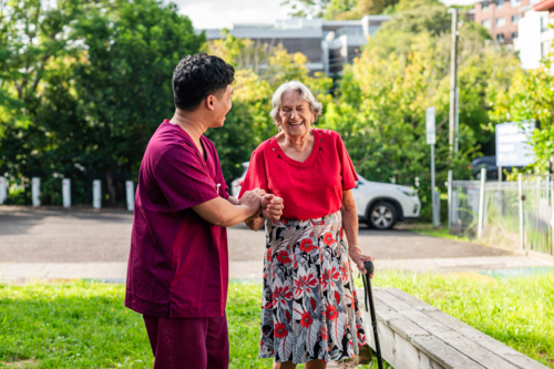 Elderly woman in a cane walking with home caregiver outdoors in grassy yard on sunny day - Australian Stock Image