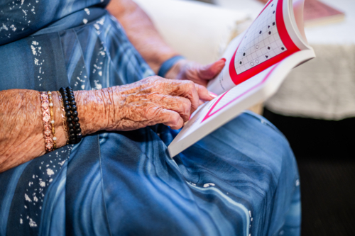 Elderly woman in blue patterned dress sitting on patterned armchair reading a book - Australian Stock Image