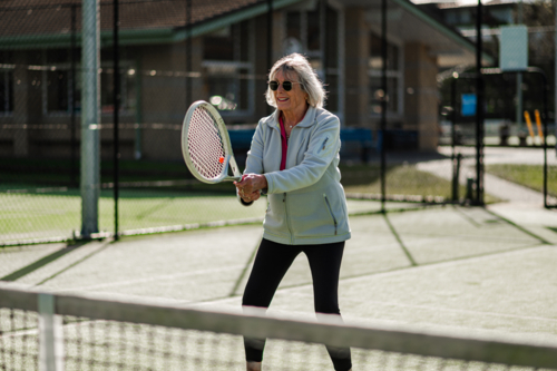 Elderly woman playing tennis, smiling and enjoying the sunny weather at an outdoor court - Australian Stock Image
