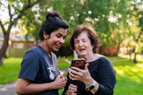 Elderly woman showing her phone to healthcare worker in dark blue scrubs outdoors - Australian Stock Image