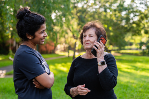 Elderly woman taking a phone call with healthcare worker stands beside her outdoors - Australian Stock Image