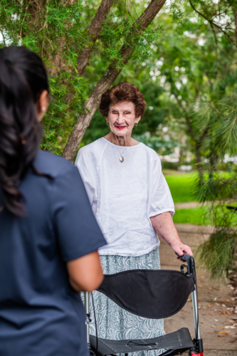 Elderly woman walking on paved footpath with her walker while care worker is approaching her - Australian Stock Image