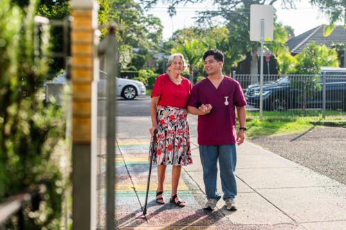 Elderly woman walking with home caregiver outdoors on paved path on sunny day - Australian Stock Image