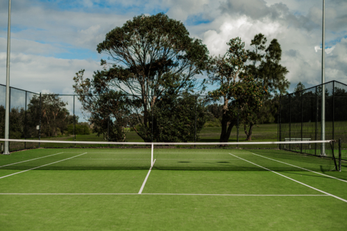 Empty tennis court features green grass and trees surrounding the area, under cloudy skies. - Australian Stock Image