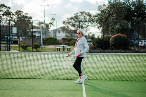 Enthusiastic woman stands by the net on an outdoor tennis court, ready to play - Australian Stock Image