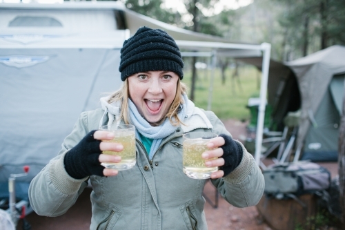 Excited young woman in beanie and gloves holding two drinks at a camp site