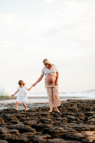 Expectant mother and daughter walking on rocky beach during sunset - Australian Stock Image