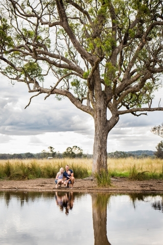 Family crouching near water of dam in paddock with large tree - Australian Stock Image