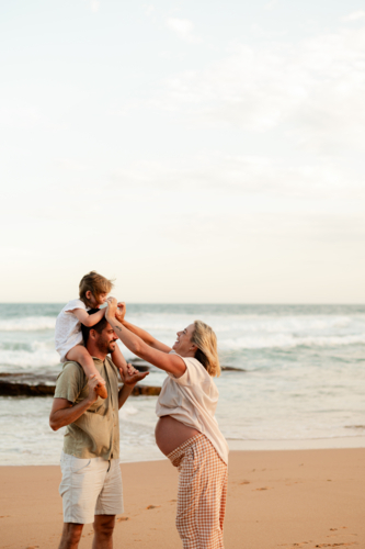 Family enjoying sunset at the beach with toddler on shoulders and pregnant woman laughing - Australian Stock Image