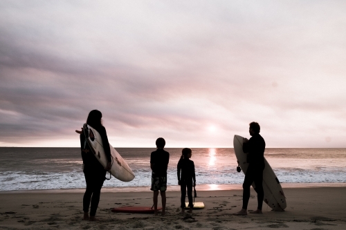 Family of four standing on beach in silhouette holding surfboards with moody sunset