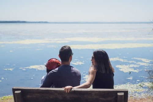 Family of three sitting on bench looking out over water