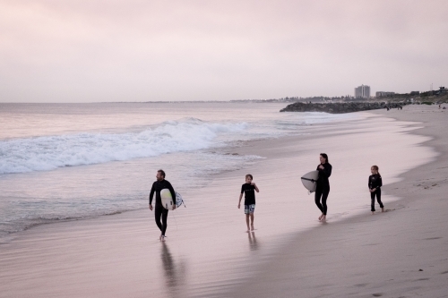 Family of two adults and two children walking along the beach carrying surfboards dressed in wetsuit