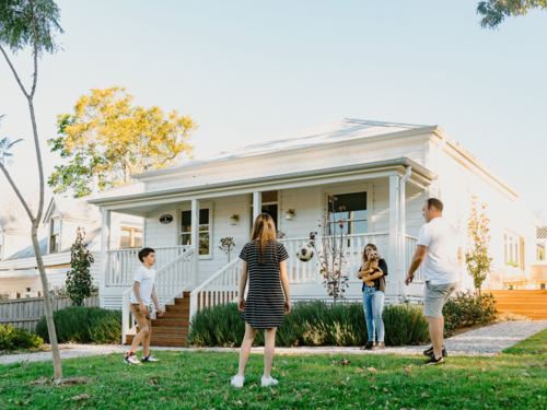 Family playing soccer in front yard of white weatherboard home in suburbs - Australian Stock Image