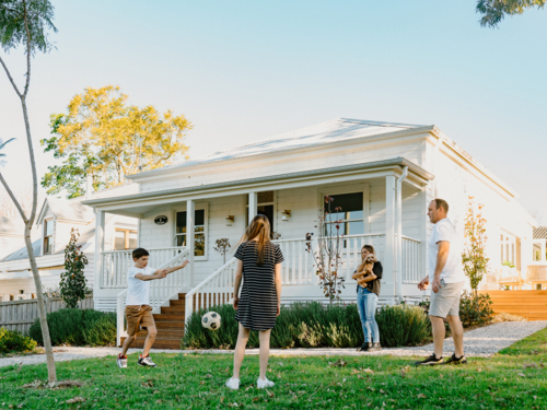 Family playing soccer in front yard of white weatherboard home in suburbs - Australian Stock Image