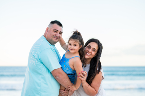 Family poses at the beach, child smiles as parents hold them with the ocean behind - Australian Stock Image