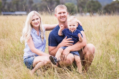 Family sitting in long grass in paddock on farm smiling - Australian Stock Image