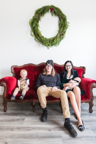 Family sitting together on red couch with Christmas wreath holding baby - Australian Stock Image