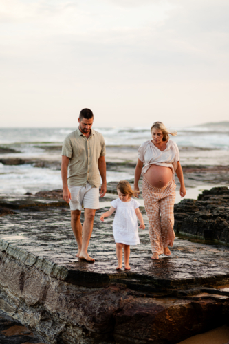 Family walking on rocky beach at sunset with pregnant mother and young daughter - Australian Stock Image