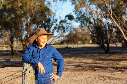 Farm kid wearing work shirt and hat leaning on a fencepost. - Australian Stock Image