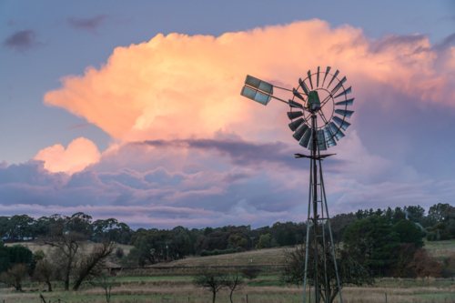 Farm windmill silhouetted against a pink thunderhead over rural farmland - Australian Stock Image