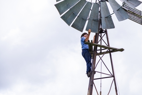 Farmer climbing up farm windmill to check and repair pump - Australian Stock Image