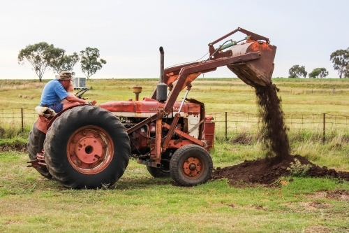 Farmer with grandchild on lap sitting on tractor emptying soil out of scoop - Australian Stock Image