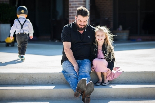 Father and daughter sitting on steps of home with young child in background - Australian Stock Image
