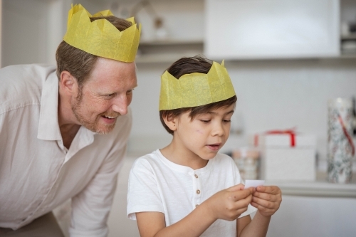 Father and son wearing yellow crowns reading Christmas cracker joke - Australian Stock Image