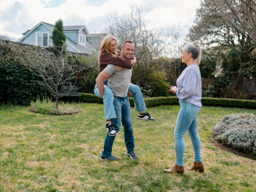 Father carrying teenage daughter piggyback while mum watches them in the garden - Australian Stock Image