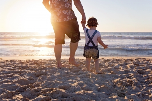 Father Holding His Daughter's Hand Walking Towards The Ocean At Sunset - Australian Stock Image