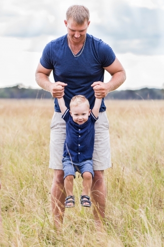 Father lifting up child by hands standing in long grass on farm - Australian Stock Image