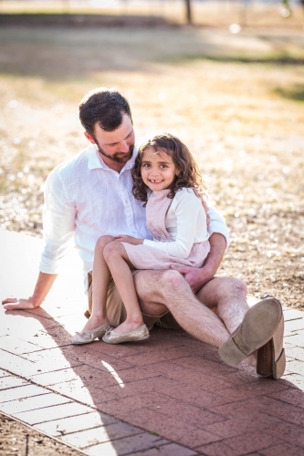 Father looking down at mixed race aboriginal and caucasian daughter sitting on lap - Australian Stock Image