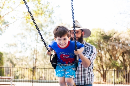 Father pushing son on the swing at the park - Australian Stock Image