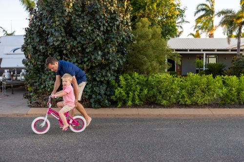 father pushing young child on two-wheeler bike on quiet street
