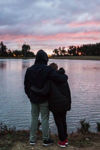 Father standing with arm around son looking out at river water at sunset - Australian Stock Image