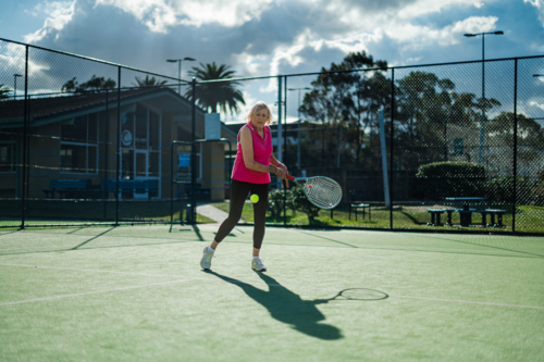 Female athlete hitting a tennis ball on a court surrounded by greenery and blue skies - Australian Stock Image
