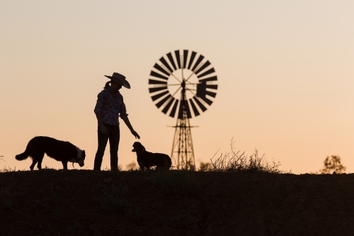 Female farmer petting dogs in silhouette with windmill - Australian Stock Image