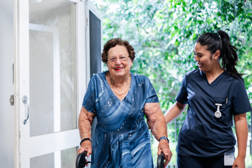 Female health caregiver supporting an elderly woman using a walker walk through the doorway - Australian Stock Image