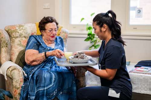 Female healthcare worker carrying a tray of snacks for an elderly woman seated on patterned armchair - Australian Stock Image