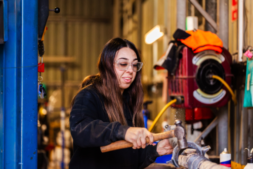 Female mechanic striking vehicle drive shaft with a hammer working on car repairs in workshop - Australian Stock Image
