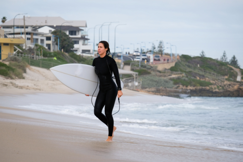 Female surfer in wetsuit walking out of ocean holding shortboard surfboard with coastal background - Australian Stock Image